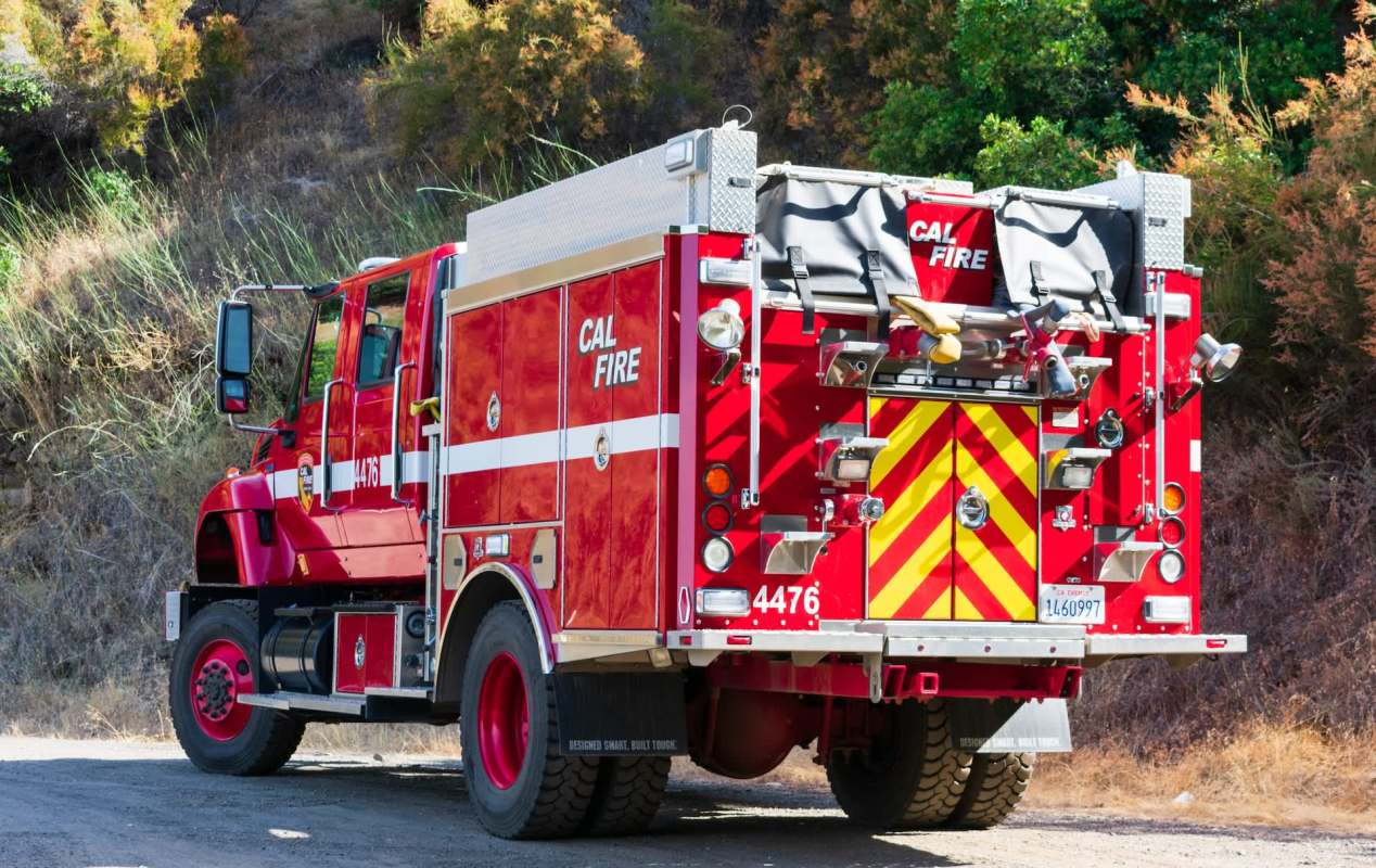 A red Cal Fire truck parked on a dirt road, next to dry vegetation on a hillside.