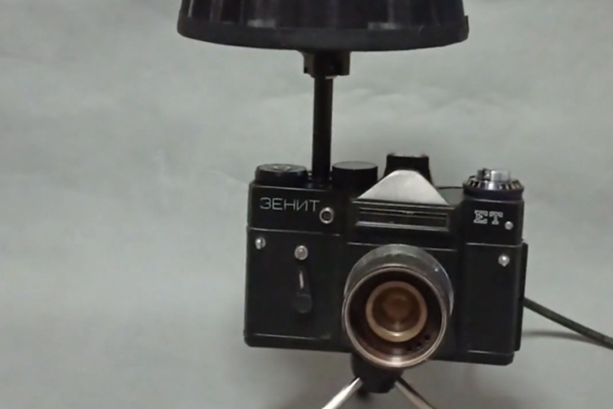 A vintage Zenit camera on a tripod with a lamp attached, set against a plain background.