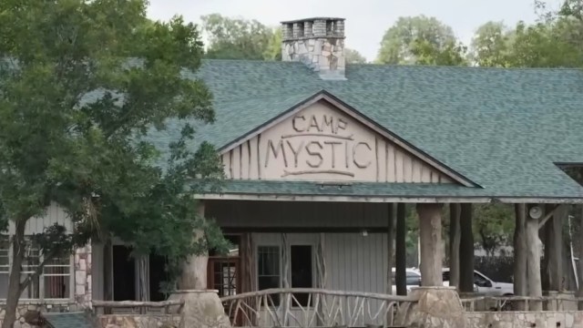 A wooden building with a green roof displays the sign "CAMP MYSTIC" surrounded by trees.