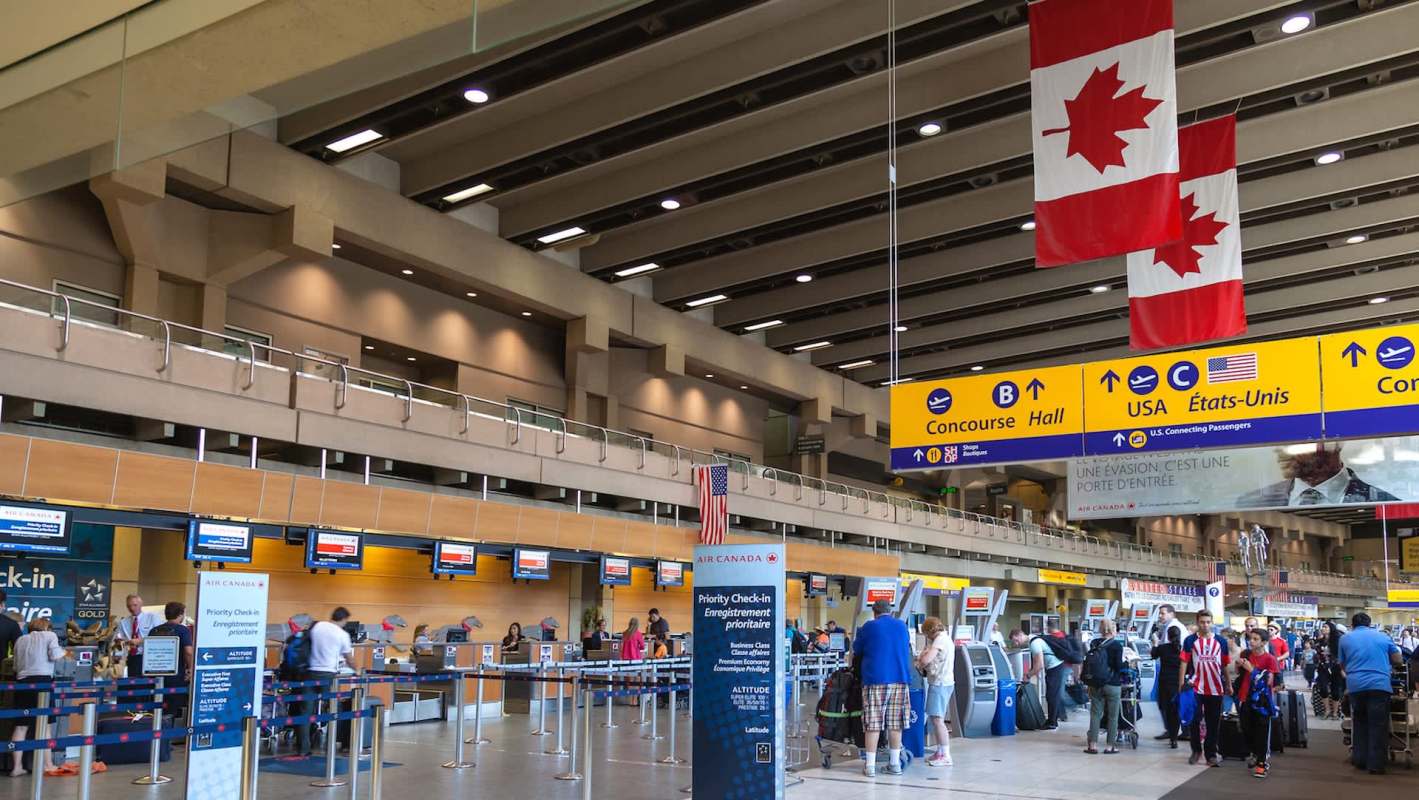 A busy airport check-in area featuring Canadian flags hanging from the ceiling, with travelers at counters and kiosks.