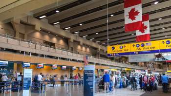 A busy airport check-in area featuring Canadian flags hanging from the ceiling, with travelers at counters and kiosks.
