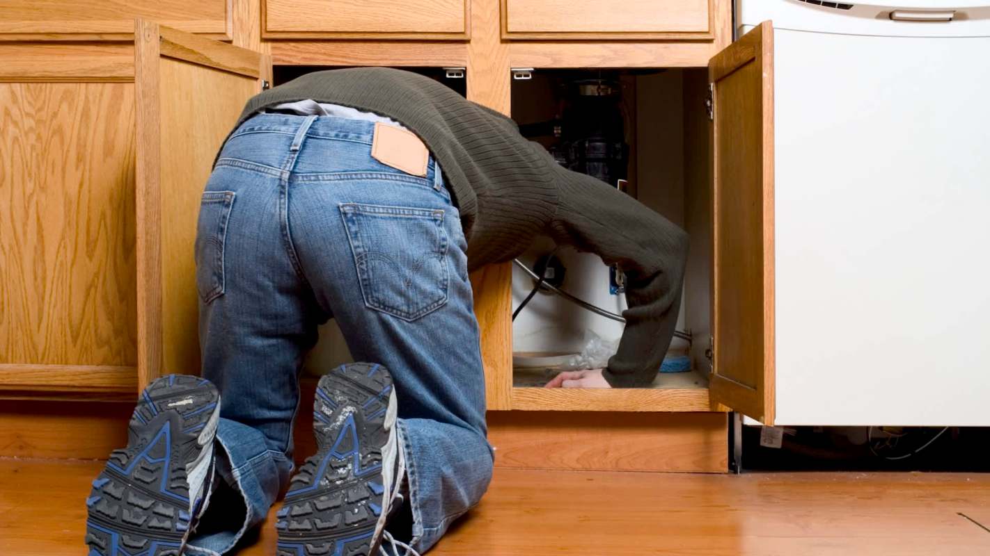 A person crouches inside a kitchen cabinet, reaching towards plumbing under the sink.