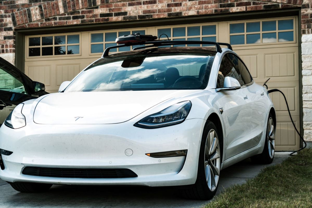 A white Tesla Model 3 charging in a driveway in front of a brick house with a brown garage.