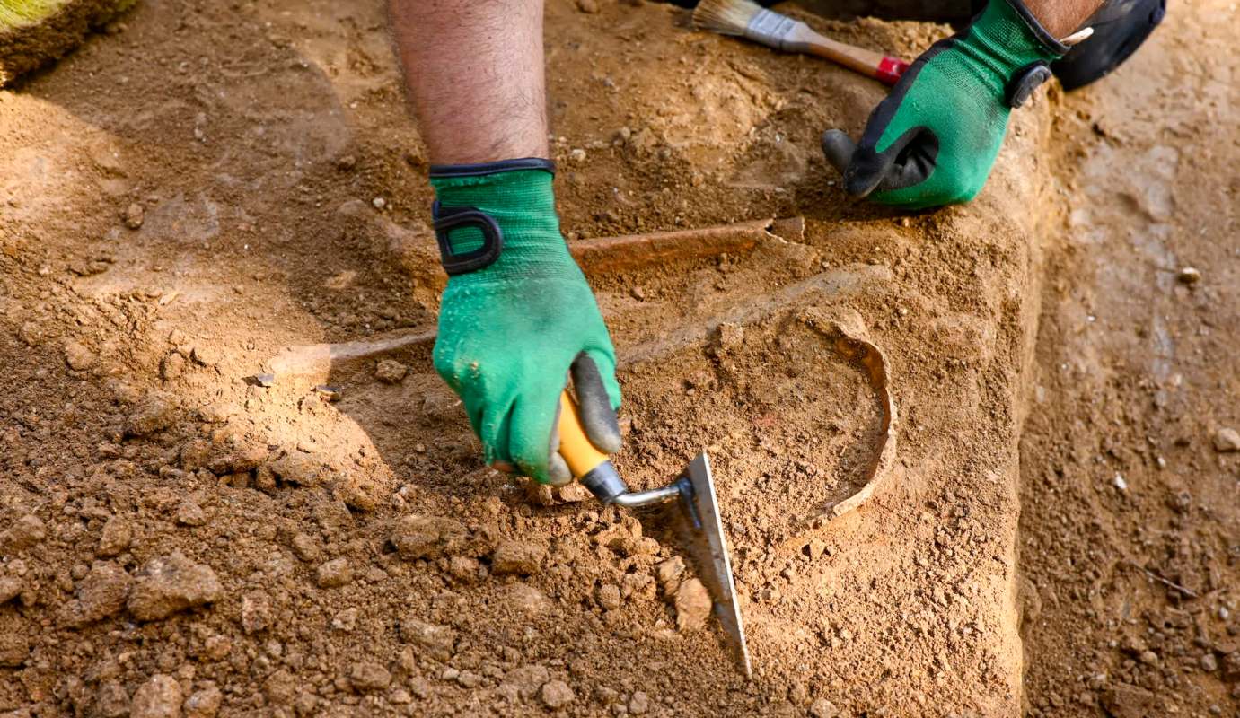 A person wearing green gloves uses a trowel to excavate soil and uncover an artifact.