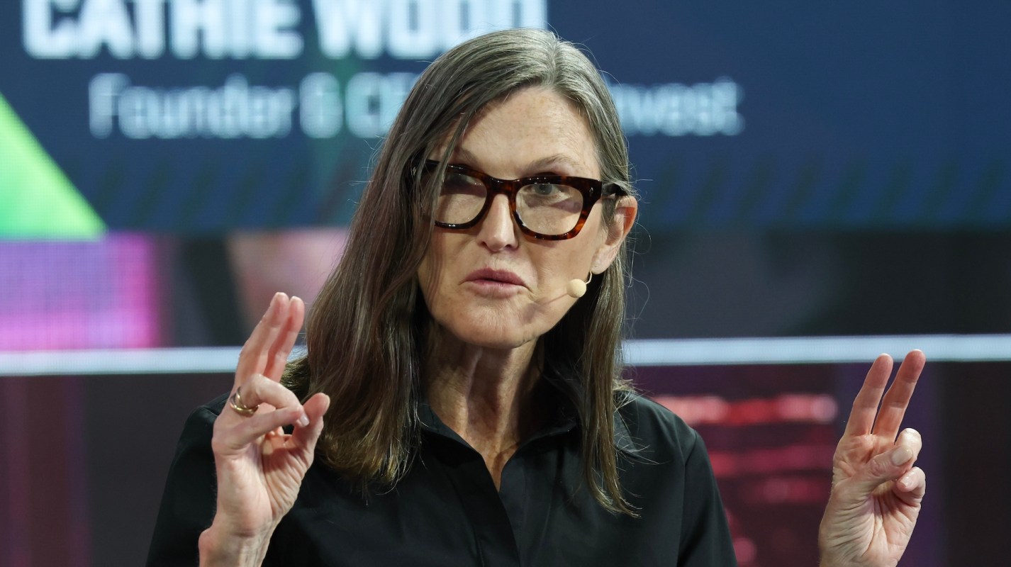 Cathie Wood gestures while speaking at a conference, with a presentation display behind her.