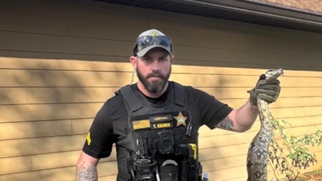 A police officer in tactical gear holds a large snake in front of a light-colored wooden building.