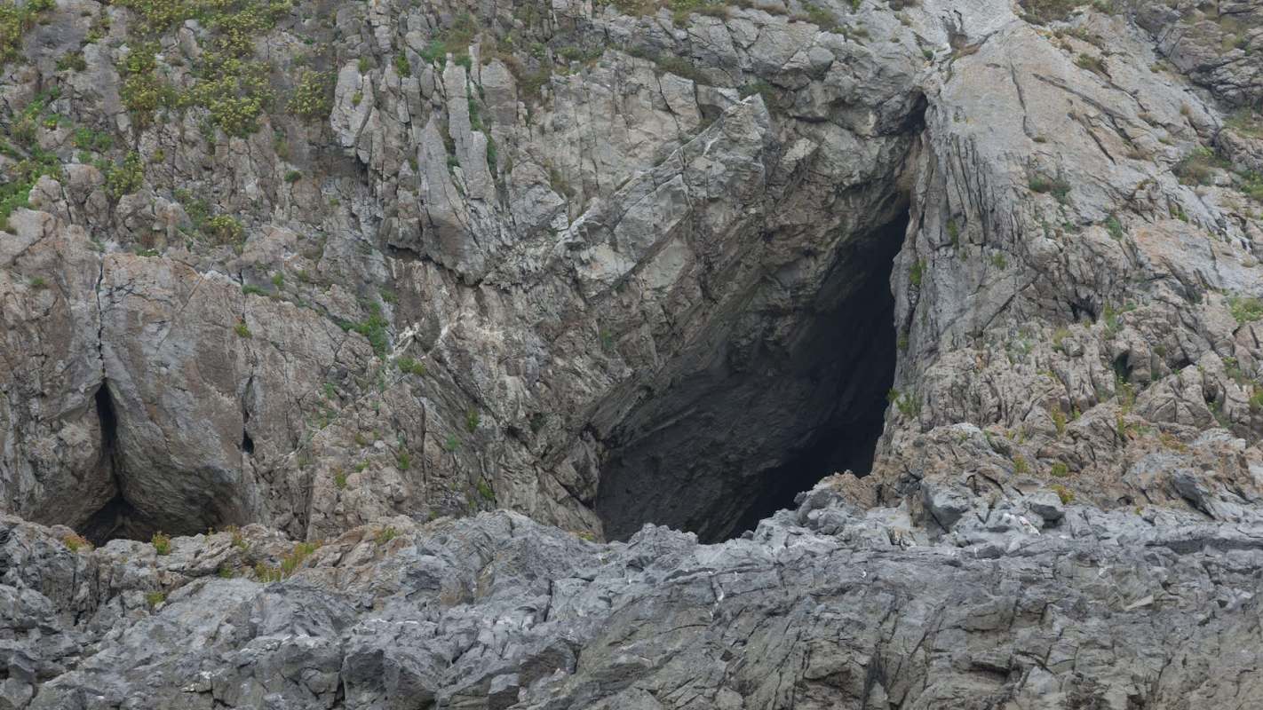 A rugged rock formation with a dark cave entrance and sparse vegetation on the surface.