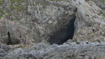 A rugged rock formation with a dark cave entrance and sparse vegetation on the surface.
