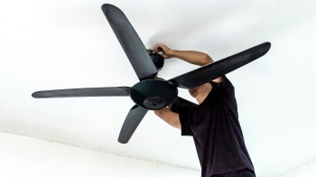 A person is repairing a black ceiling fan, reaching up toward the fan blades.