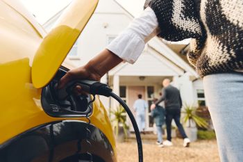 A person plugs in a charging cable to a yellow electric car while family members walk toward a house in the background.