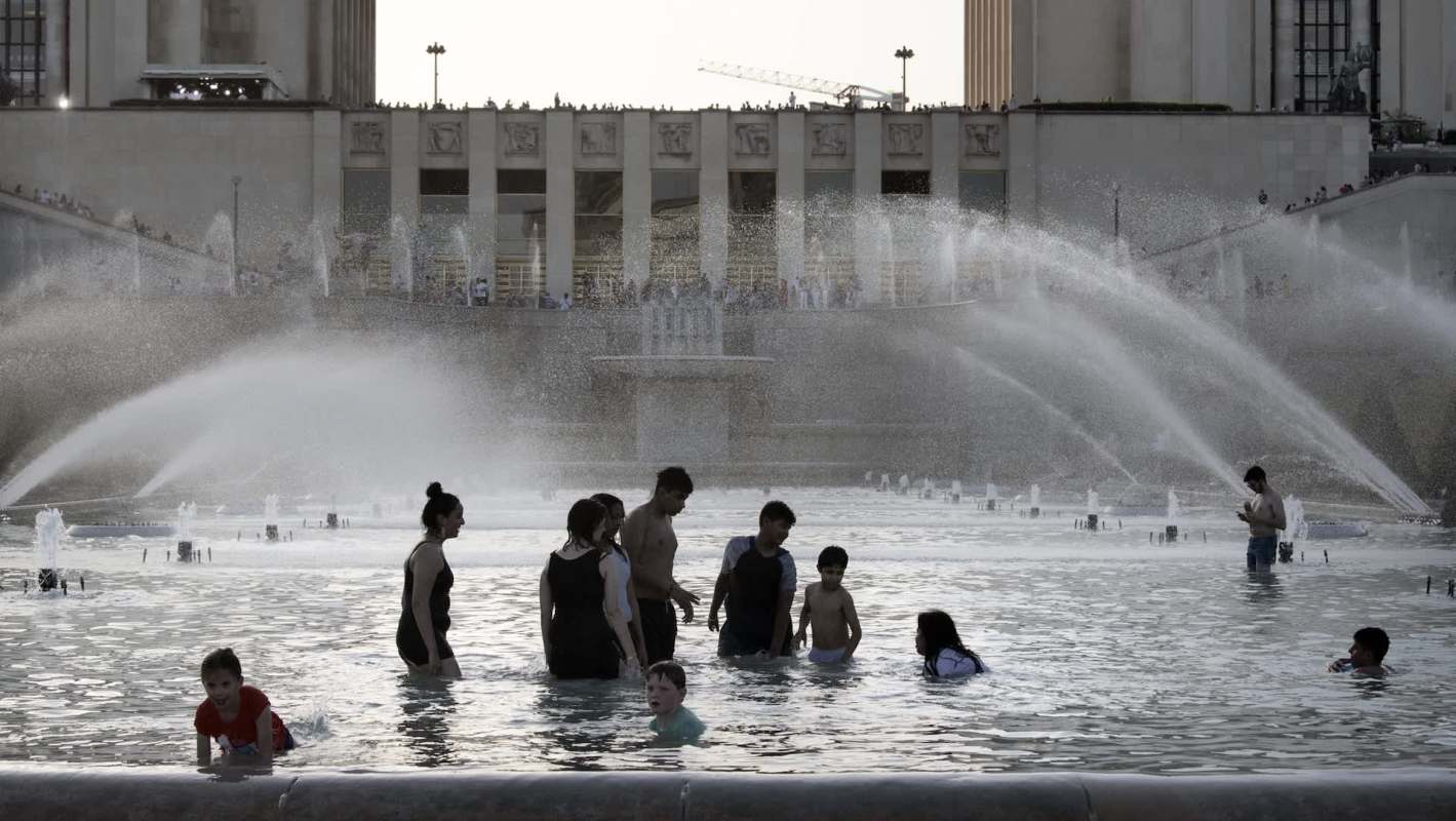 People playing and splashing in a fountain pool, with sprays of water in front of a large building in the background.