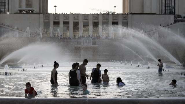 People playing and splashing in a fountain pool, with sprays of water in front of a large building in the background.