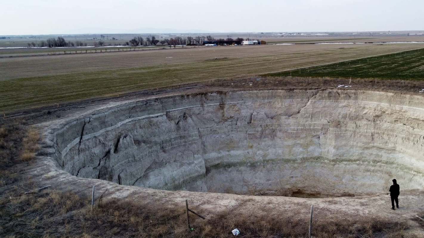 A person stands at the edge of a large, deep sinkhole in a rural landscape.
