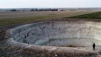 A person stands at the edge of a large, deep sinkhole in a rural landscape.