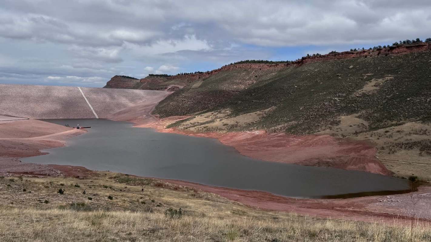 A reservoir surrounded by reddish hills under a cloudy sky, with a small boat near the shore.