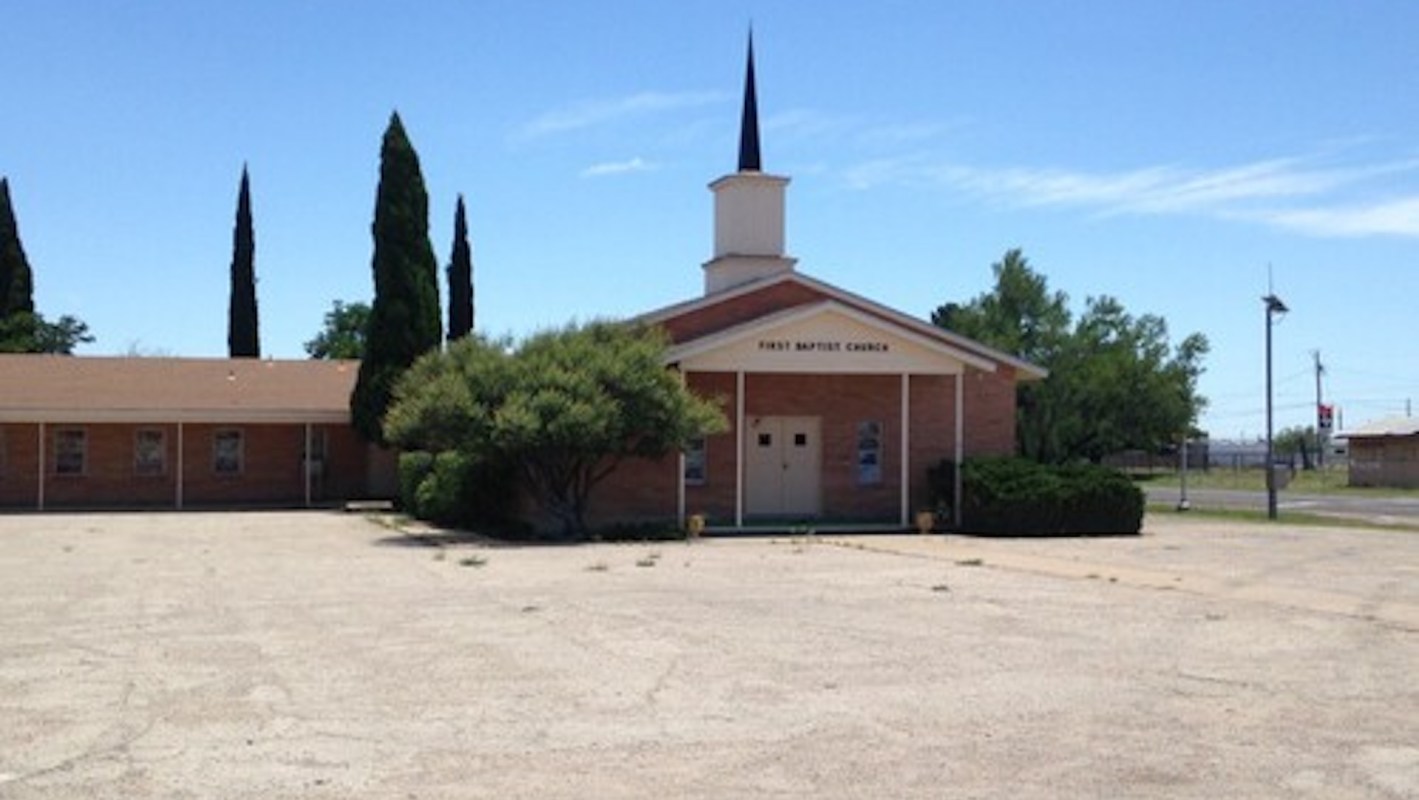 A brick building with a steeple, labeled "First Baptist Church," surrounded by trees and sparse vegetation.