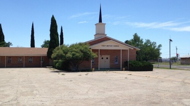 A brick building with a steeple, labeled "First Baptist Church," surrounded by trees and sparse vegetation.