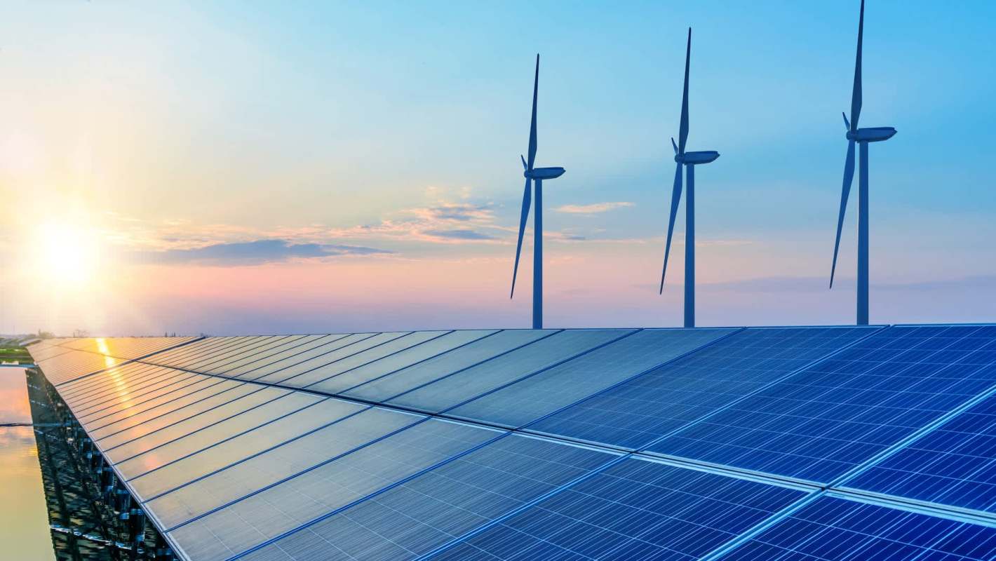 Solar panels in the foreground with wind turbines and a sunset sky in the background.