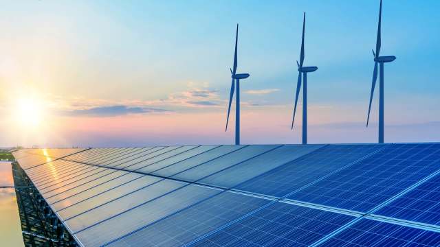 Solar panels in the foreground with wind turbines and a sunset sky in the background.