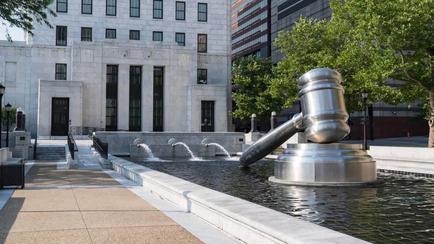A large metallic gavel sculpture stands above a fountain in front of a modern building surrounded by greenery.