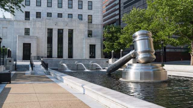 A large metallic gavel sculpture stands above a fountain in front of a modern building surrounded by greenery.