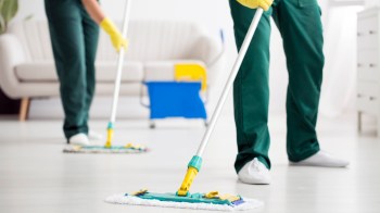 Two individuals in green uniforms use mops to clean a bright, modern room with a couch and cleaning supplies nearby.