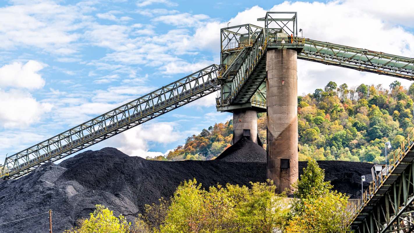 A coal conveyor system transports material over a large mound of coal against a backdrop of trees and blue skies.