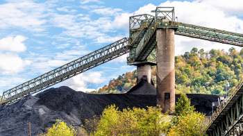 A coal conveyor system transports material over a large mound of coal against a backdrop of trees and blue skies.