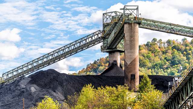 A coal conveyor system transports material over a large mound of coal against a backdrop of trees and blue skies.