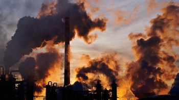 A silhouette of industrial smokestacks emitting dark smoke against a vibrant sunset sky.