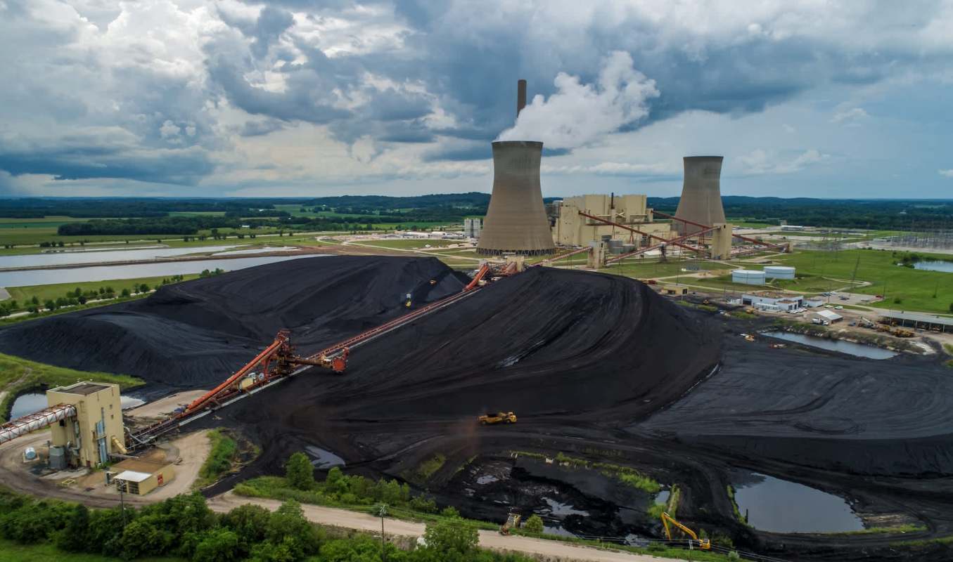 A coal storage facility with large mounds of coal and cooling towers under a cloudy sky.