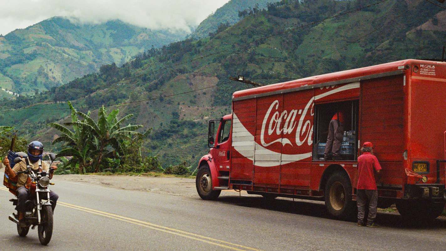 A Coca-Cola delivery truck stopped on a rural road with a motorcycle rider passing by in a mountainous landscape.