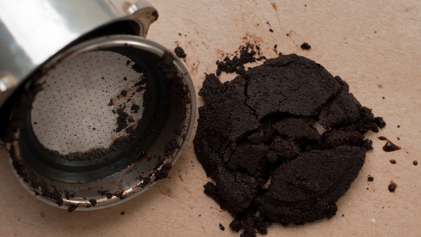 A spilled coffee grounds puck next to a metallic coffee filter on a light beige surface.
