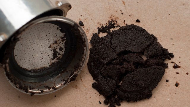 A spilled coffee grounds puck next to a metallic coffee filter on a light beige surface.