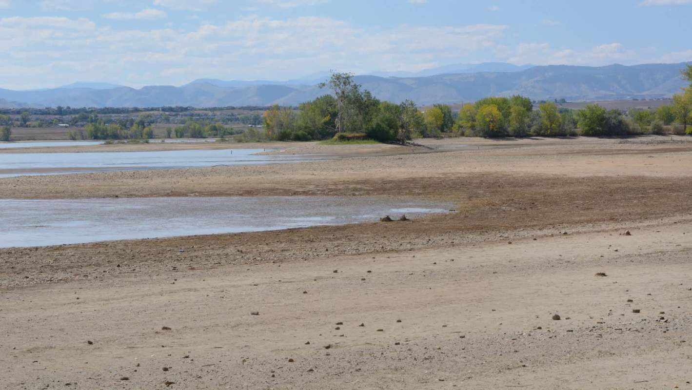 A barren shoreline with sparse vegetation, revealing low water levels and distant mountains under a partly cloudy sky.