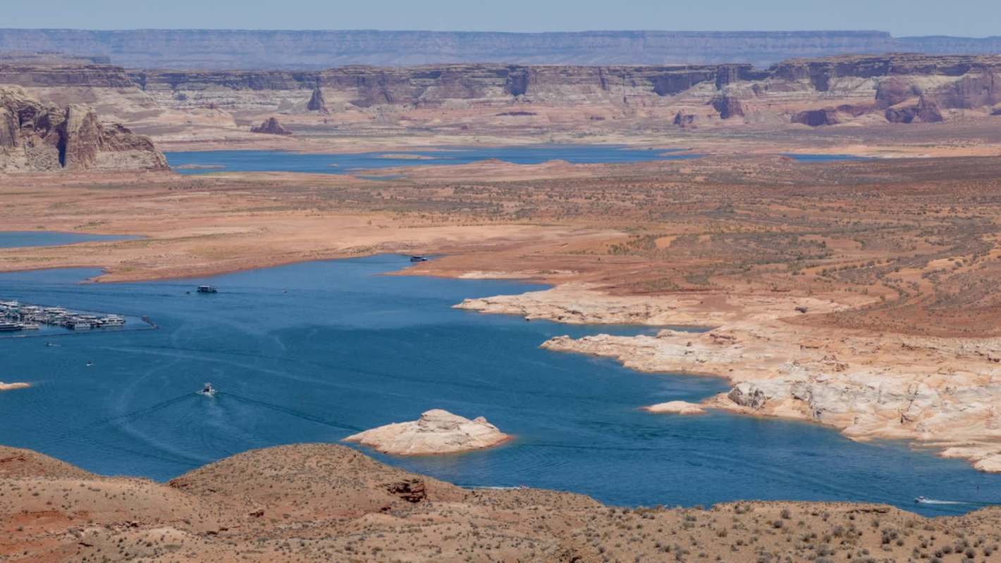 A panoramic view of a lake surrounded by arid, rocky terrain and distant cliffs under clear blue skies.