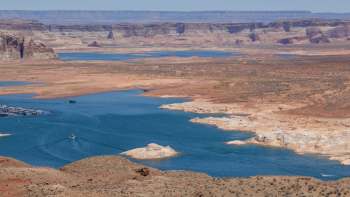 A panoramic view of a lake surrounded by arid, rocky terrain and distant cliffs under clear blue skies.
