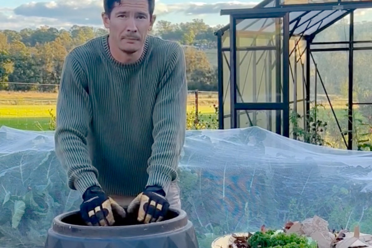A person wearing a sweater and gloves is mixing soil in a large compost barrel in a garden setting.
