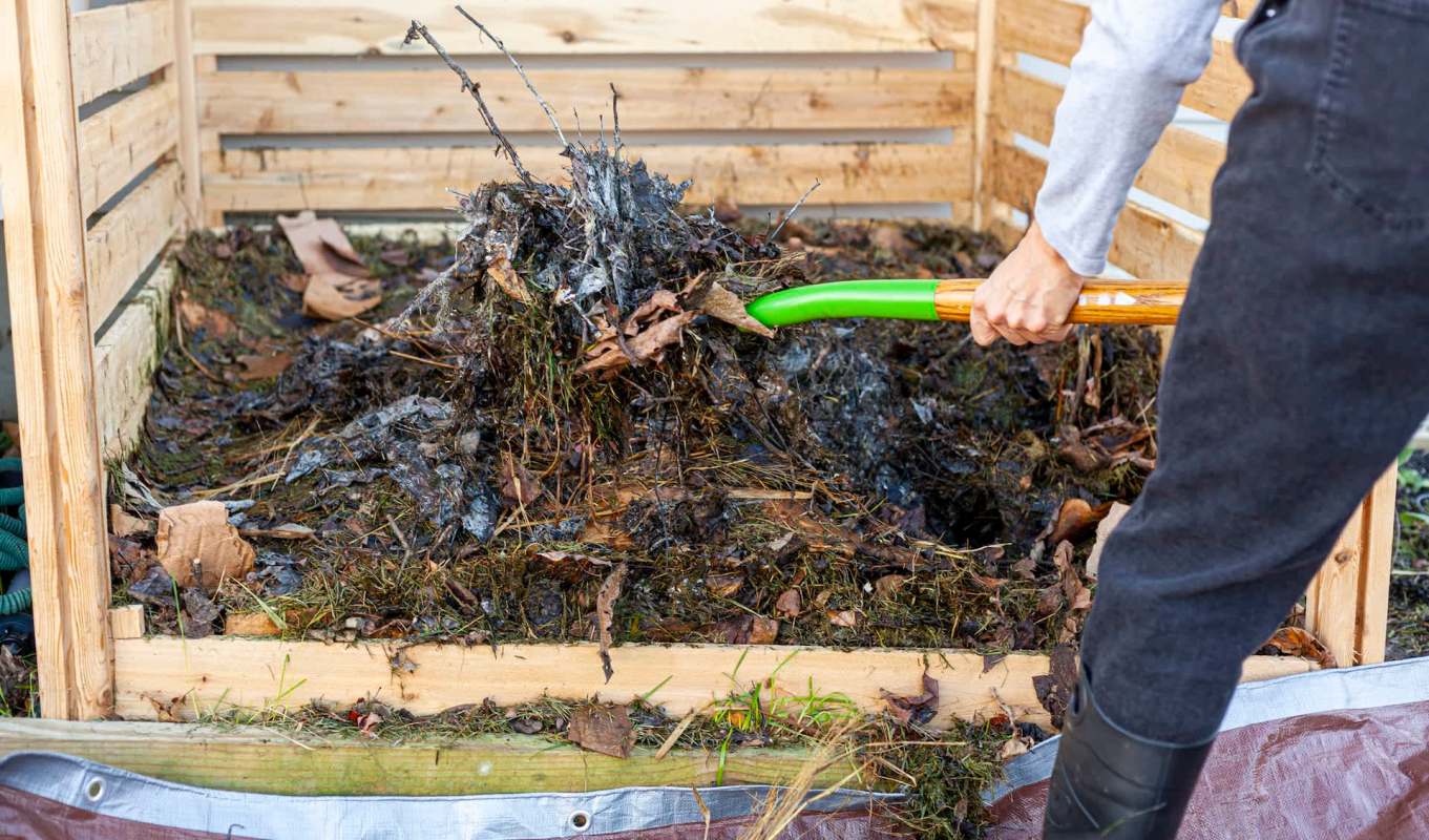 A person uses a green shovel to turn compost in a wooden bin filled with organic materials and leaves.