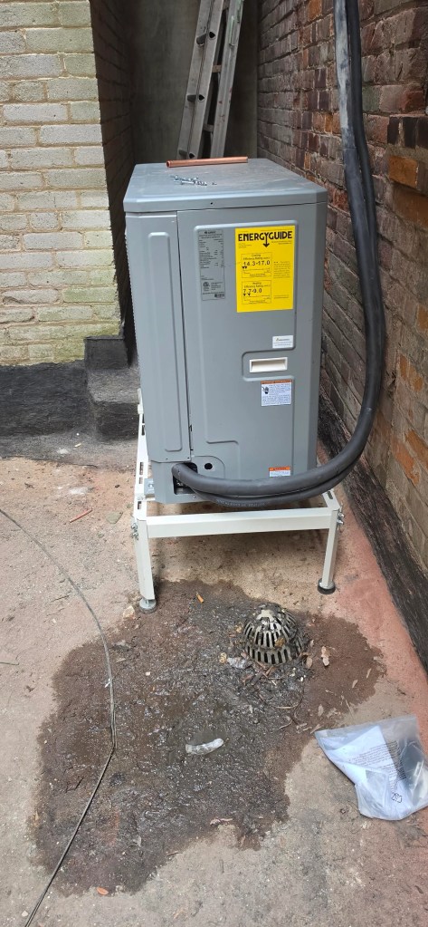 An HVAC unit on a snow stand near a brick wall. 