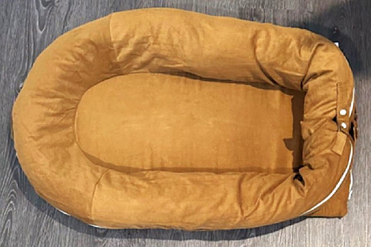 A tan, plush baby lounger bed with raised edges, placed on a dark wooden floor.