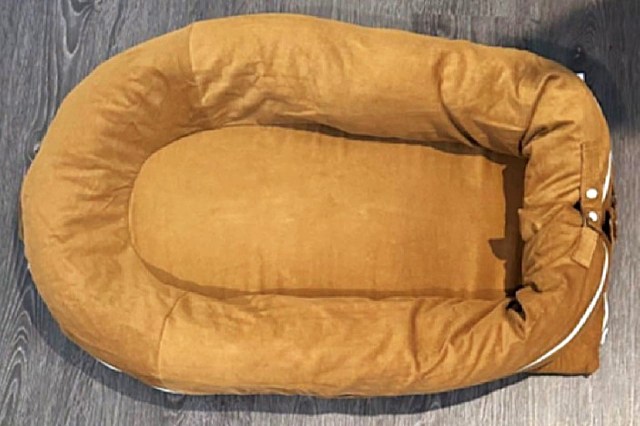 A tan, plush baby lounger bed with raised edges, placed on a dark wooden floor.