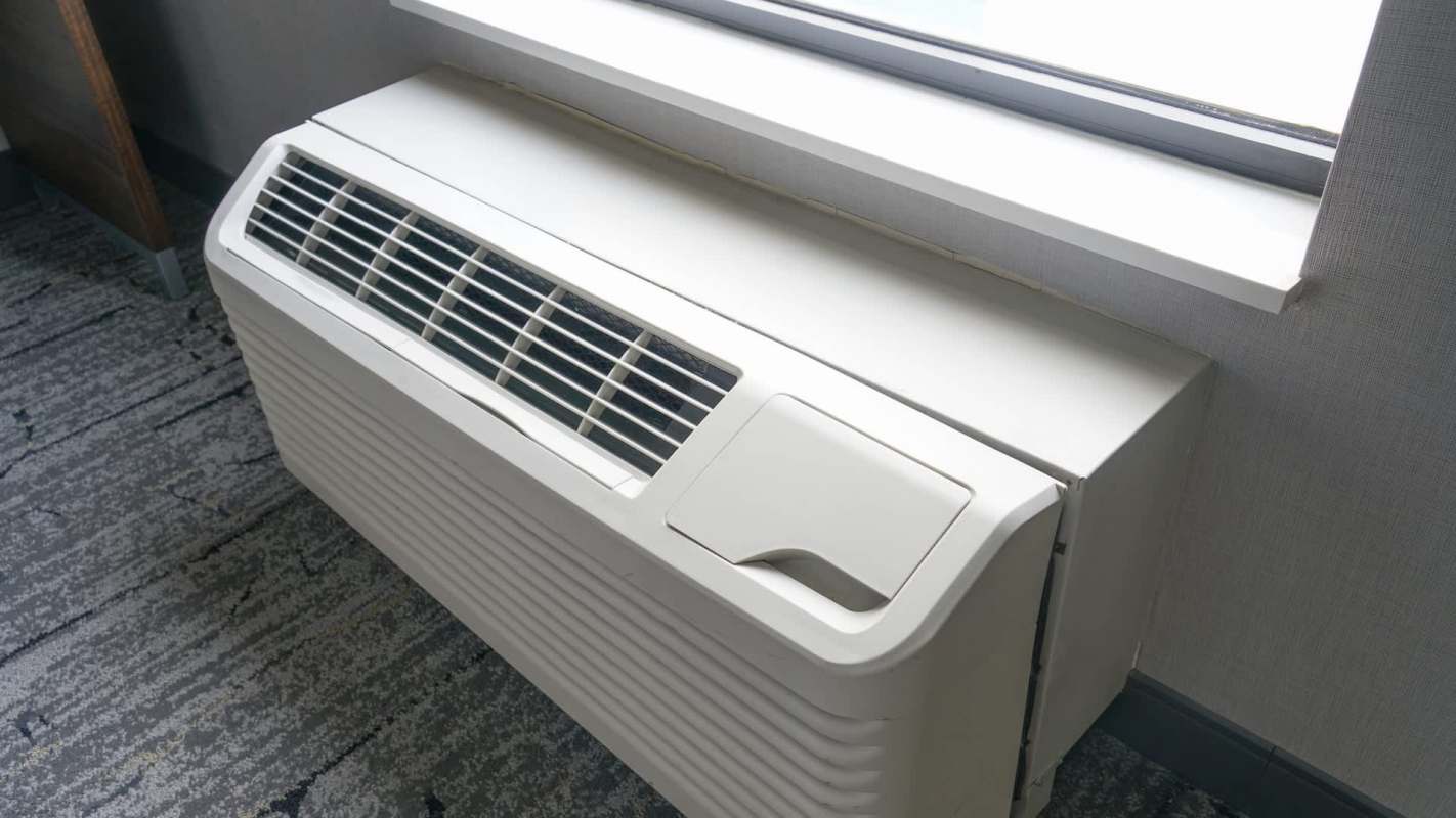 A white air conditioning unit installed beneath a window in a room with gray carpeting.