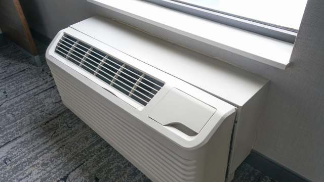 A white air conditioning unit installed beneath a window in a room with gray carpeting.