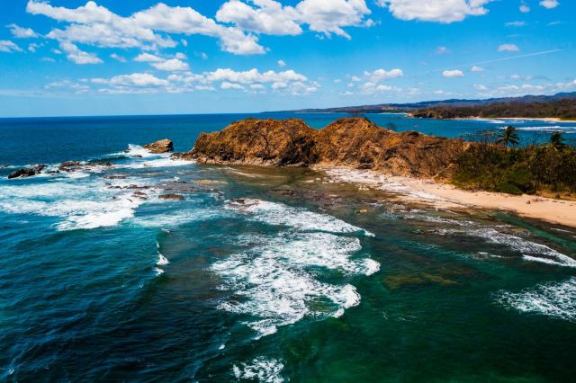 Aerial view of rocky coastline with turquoise water and sandy beaches under a bright blue sky with scattered clouds.