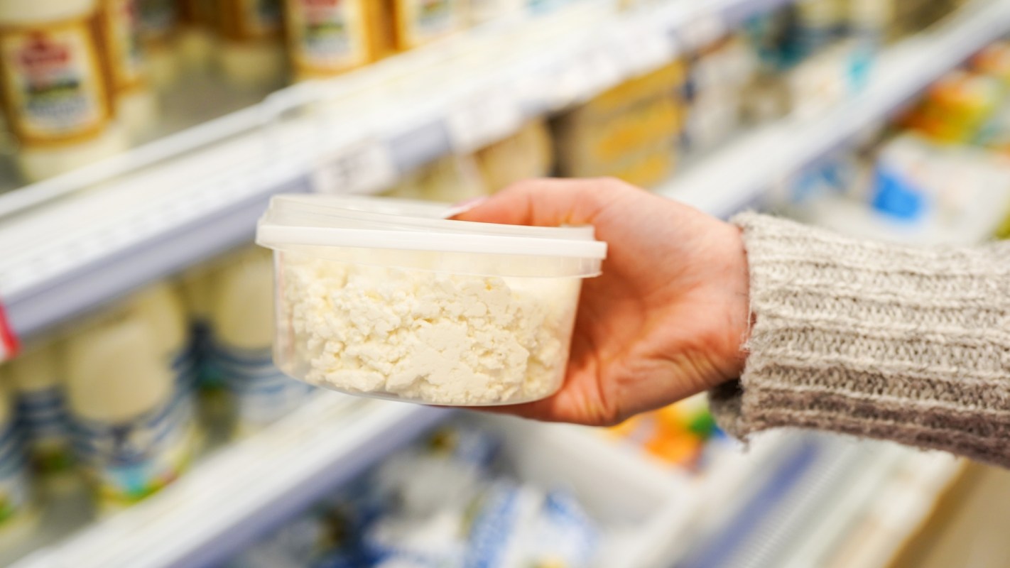 A hand holding a plastic container of cottage cheese in a grocery store aisle.