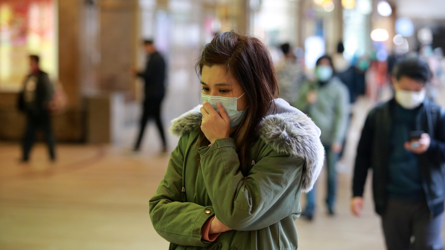 A young woman in a green coat and mask stands pensively in a busy indoor space while others pass by.