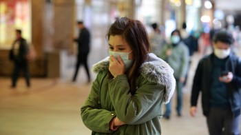 A young woman in a green coat and mask stands pensively in a busy indoor space while others pass by.