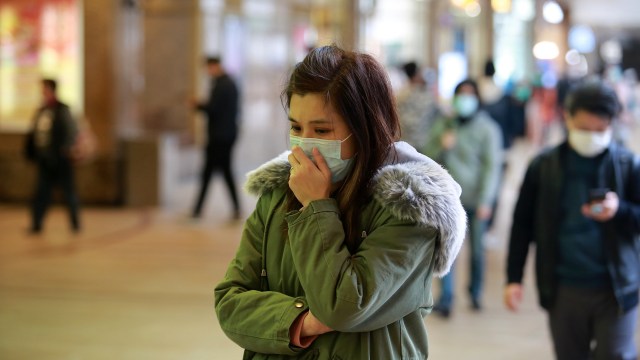 A young woman in a green coat and mask stands pensively in a busy indoor space while others pass by.