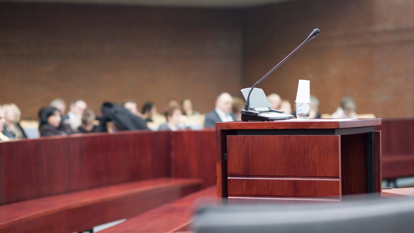 A courtroom setting with a wooden podium, microphone, and a blurred audience in the background.